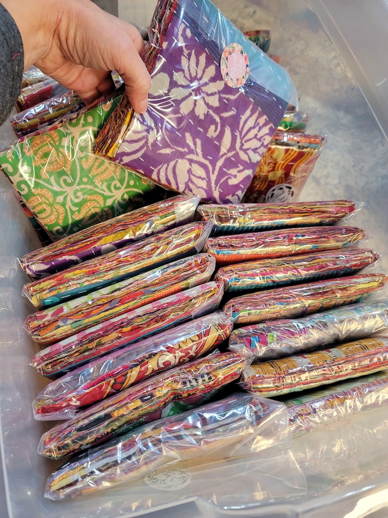 May include: A stack of colorful fabric squares wrapped in plastic, some with floral patterns, others with geometric designs. The fabric squares are arranged in a clear plastic container.