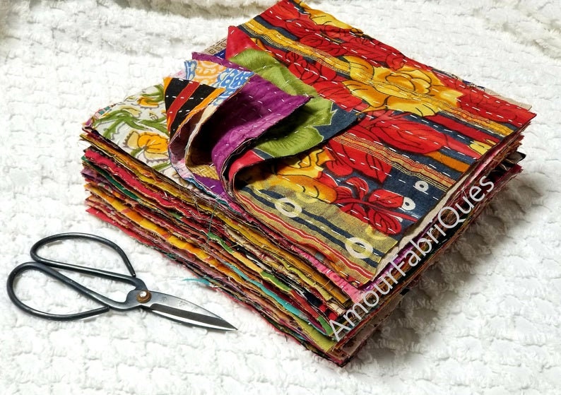 May include: A stack of colorful fabric squares, each with a different pattern, are neatly arranged. The fabric squares are made of a variety of materials, including cotton and silk. The squares are all about 12 inches by 12 inches in size. A pair of black scissors is lying on a white surface in the foreground.