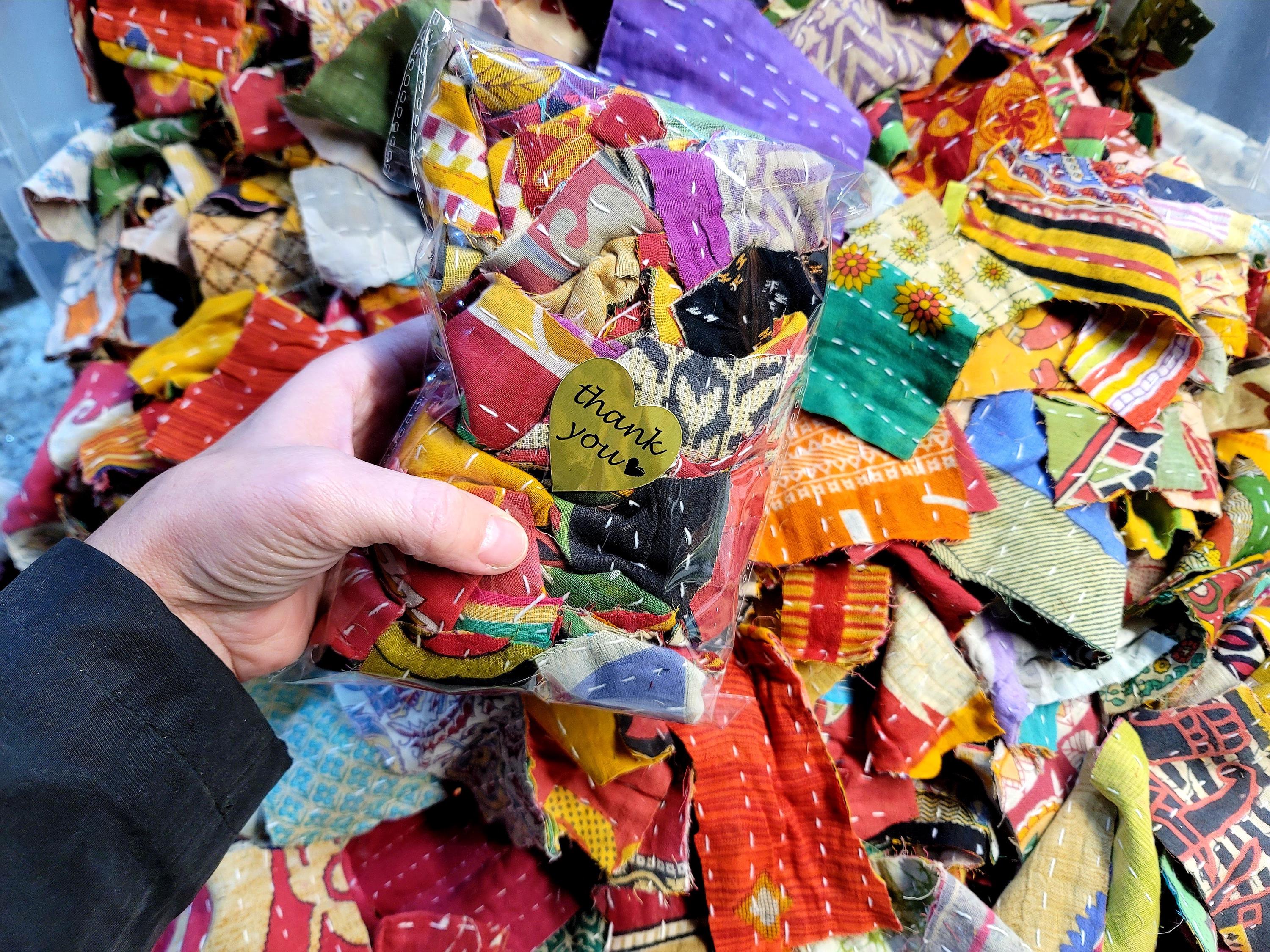 May include: A plastic bag filled with colorful fabric scraps, some of which are stitched together. The bag is held in a hand, and a small gold heart-shaped tag with the text "thank you" is attached to the bag.
