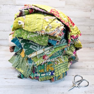 May include: A pile of assorted green fabric scraps with white stitching, featuring floral and geometric patterns. The text "Amour Fabriques" is visible on some pieces. A pair of silver scissors rests on the wooden surface.
