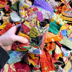 Könnte beinhalten: Ein Plastikbeutel gefüllt mit bunten Stoffresten, von denen einige zusammengenäht sind. Der Beutel wird in einer Hand gehalten, und ein kleines goldenes herzförmiges Etikett mit dem Text "Danke" ist am Beutel befestigt.