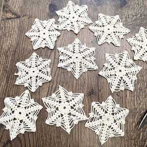 May include: A collection of eleven cream-colored, crocheted snowflake ornaments arranged on a wooden surface. The snowflakes have intricate, openwork designs. A pair of black scissors is visible in the lower right corner.