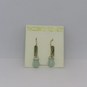 May include: A pair of gold dangle earrings with a light blue teardrop-shaped stone and white pearl accents. The earrings are on a white card with the text "THOUSAND FLOWERS" and "PRECIOUS S".