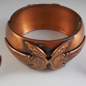 Puede incluir: Un conjunto de joyas de cobre que incluye un brazalete ancho y dos pendientes a juego. El brazalete y los pendientes tienen un diseño decorativo de alas de mariposa estilizadas.