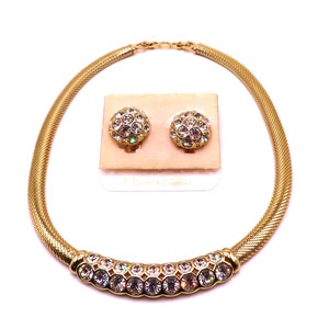 May include: A gold tone necklace and matching earrings set with clear crystal stones. The necklace has a mesh chain and a curved row of stones. The earrings are clip-on style.