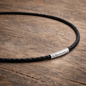 Può includere: Una collana in pelle intrecciata nera con chiusura argentata. La collana è esposta su una superficie di legno. La chiusura è di colore argento lucido e ha una chiusura magnetica. La pelle è finemente intrecciata.
