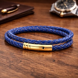 May include: A blue braided leather bracelet with a gold clasp. The bracelet is wrapped twice and rests on a wooden surface. A watch and wallet are in the background.