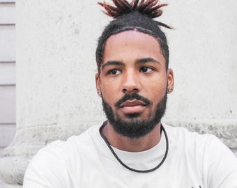 Pode incluir: Um homem com dreadlocks e barba está vestindo uma camiseta branca com um colar trançado preto.