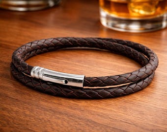 Puede incluir: Una pulsera de cuero marrón con un diseño trenzado. La pulsera se envuelve dos veces y presenta un cierre plateado. La pulsera se exhibe sobre una superficie de madera.
