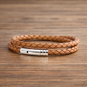 Op de afbeelding: Een lichtbruine, gevlochten leren armband met een zilveren magneetsluiting. De armband is twee keer gewikkeld en ligt op een houten oppervlak. Het leer heeft een getextureerde uitstraling.