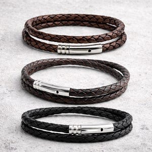 May include: Three braided leather bracelets in shades of brown and black, each with a silver-colored clasp. The bracelets are arranged in a stack, showcasing the different color options and the texture of the woven leather. The bracelets are designed to wrap around the wrist.