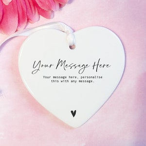 May include: White ceramic heart-shaped ornament with a white ribbon. The ornament has black text that reads "Your Message Here" and "Your message here, personalise this with any message." There is a small black heart at the bottom of the ornament.