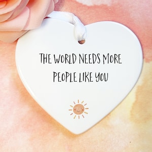 May include: A white heart-shaped ceramic ornament featuring the black text THE WORLD NEEDS MORE PEOPLE LIKE YOU and a small illustration of a smiling sun. A white ribbon is threaded through a hole at the top for hanging purposes.
