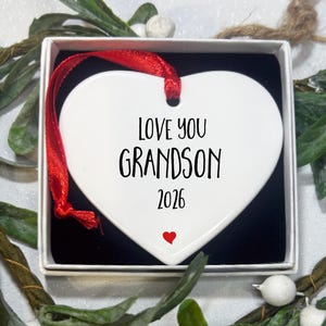 May include: A white heart-shaped ceramic ornament featuring the black text LOVE YOU GRANDSON 2026 and a small red heart icon. The ornament is suspended by a red ribbon and is presented inside a square white gift box with a black interior lining.