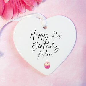 May include: White ceramic heart-shaped ornament with a white ribbon. The ornament has black text that reads "Happy 21st Birthday Katie" and a pink cupcake illustration.