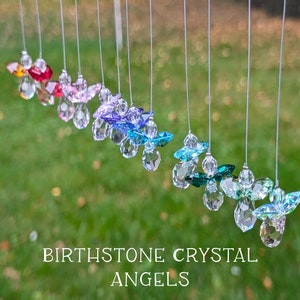 May include: A rainbow of crystal angel ornaments hanging from white strings against a green grass background. The text "Birthstone Crystal Angels" is displayed below the ornaments.