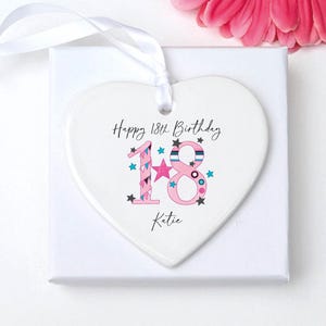 May include: A white heart-shaped ornament with the text "Happy 18th Birthday Katie." The number "18" is pink and decorated with stars. A white ribbon is attached for hanging. The ornament is on a white box.