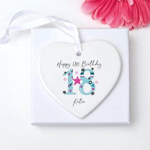May include: A white heart-shaped ornament with the text "Happy 18th Birthday Katie." The number 18 is decorated with blue, pink, and teal accents. A white ribbon is attached for hanging. A pink flower is in the background.