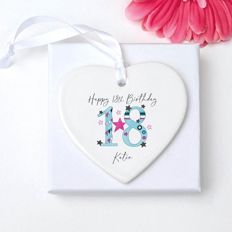 May include: A white heart-shaped ornament with the text "Happy 18th Birthday Katie." The number 18 is decorated with blue, pink, and teal accents. A white ribbon is attached for hanging. A pink flower is in the background.