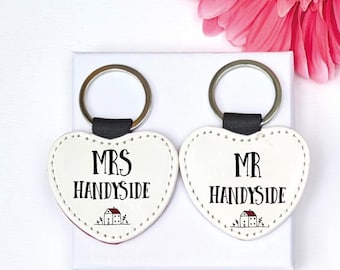 May include: Two heart-shaped keychains with the text "MRS HANDYSIDE" and "MR HANDYSIDE" in black. Each keychain has a small house graphic. The keychains are white with a black strap and silver ring.