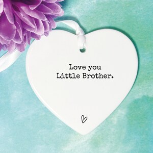May include: White ceramic heart-shaped ornament with a white ribbon. The ornament has black text that reads "Love you Little Brother." There is a small heart drawn at the bottom of the ornament.