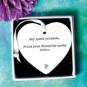 May include: White ceramic heart-shaped ornament with a ribbon. The ornament has the text "Any quote printed... Print your favourite quote here..." and a small heart symbol.