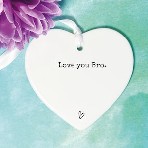 May include: White ceramic heart-shaped ornament with a white ribbon and the text "Love you Bro." in black lettering. A small heart is drawn at the bottom of the ornament.
