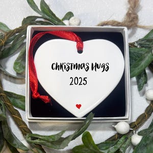 May include: A white heart-shaped Christmas ornament with the words "Christmas Hugs" and the year "2025" in black script. A small red heart is at the bottom. The ornament has a red ribbon and is in a white box with a black interior.