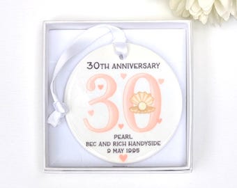 May include: A white ceramic 30th anniversary ornament with a peach-colored "30" and a pearl design. The ornament includes the text "30TH ANNIVERSARY", "PEARL", and names and a date. It is presented in a white gift box with a white ribbon.