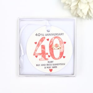 May include: A white ceramic 40th anniversary ornament in a white box. The ornament has the number "40" in coral, a ring illustration, and the text "40th Anniversary", "Ruby", names and date.
