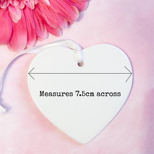 May include: A white heart-shaped ceramic ornament with a smooth, glossy finish. A white ribbon is threaded through a hole at the top for hanging. A black graphic overlay shows a horizontal arrow and the text Measures 7.5cm across.