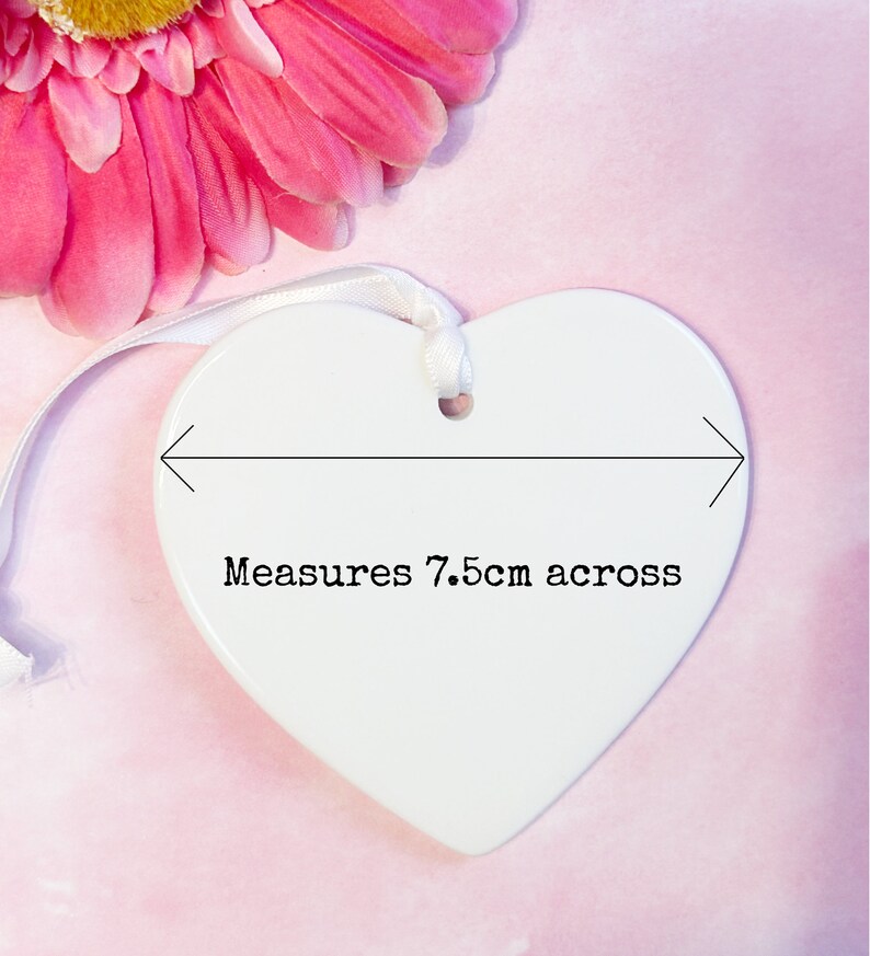 May include: A white heart-shaped ceramic ornament with a smooth, glossy finish. A white ribbon is threaded through a hole at the top for hanging. A black graphic overlay shows a horizontal arrow and the text Measures 7.5cm across.