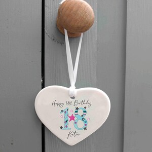 May include: A heart-shaped ceramic ornament with the text "Happy 18th Birthday Katie." The number 18 is in blue with star and pink accents. A white ribbon is attached for hanging.