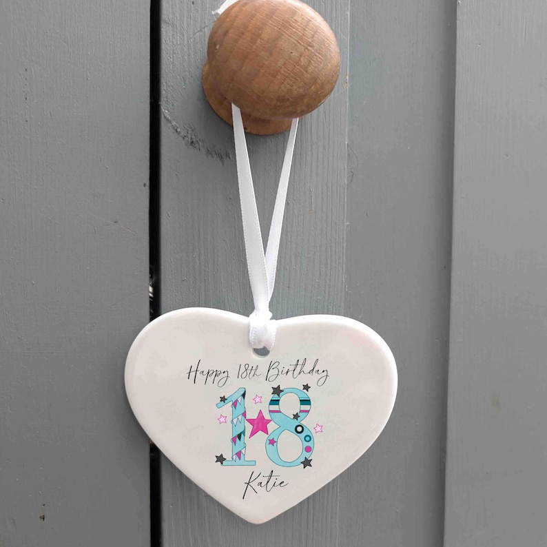 May include: A heart-shaped ceramic ornament with the text "Happy 18th Birthday Katie." The number 18 is in blue with star and pink accents. A white ribbon is attached for hanging.