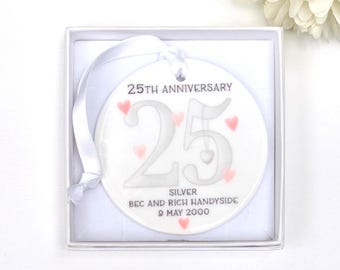 May include: A white ceramic 25th anniversary ornament with a silver "25" and pink hearts. The ornament includes the text "25th Anniversary", "Silver", and names and a date. It is presented in a white gift box with a white ribbon.