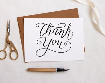 Puede incluir: Una tarjeta blanca con las palabras "Thank You" en elegante escritura negra, un sobre marrón, unas tijeras doradas y un bolígrafo dorado con una pluma negra están dispuestos sobre una superficie blanca. También hay una cinta color crema.