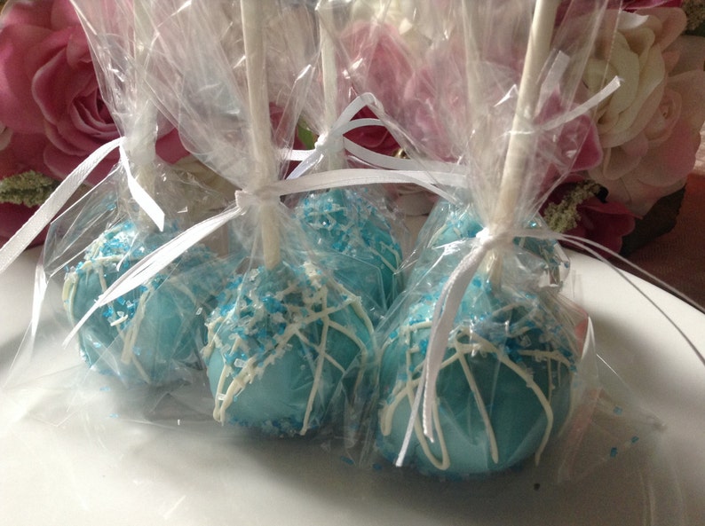 May include: Four blue and white cake pops are individually wrapped in clear cellophane bags tied with white ribbon. The cake pops are decorated with white chocolate and blue sprinkles.