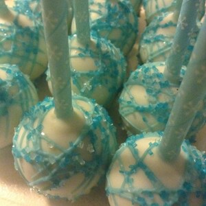 May include: White cake pops decorated with blue sprinkles and a blue icing design. The cake pops are on sticks.