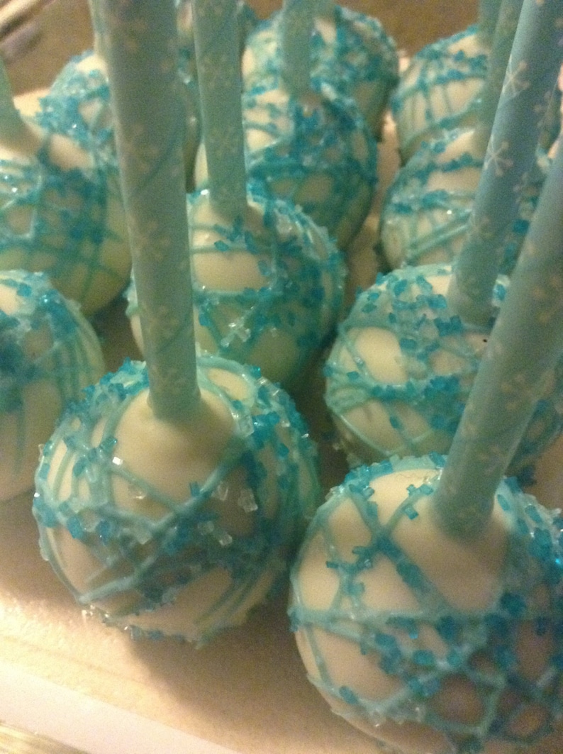 May include: White cake pops decorated with blue sprinkles and a blue icing design. The cake pops are on sticks.