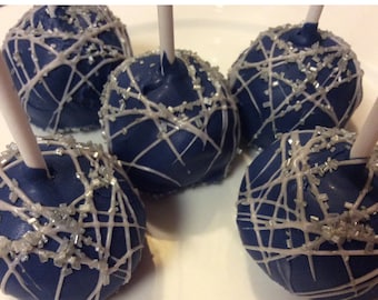 May include: Five navy blue cake pops with white drizzle and silver sprinkles. Each cake pop is round and sits on a white stick. The white drizzle creates a web-like pattern across the surface of the cake pops.