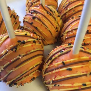 May include: Close-up of several orange cake pops with white sticks. The cake pops are decorated with brown and yellow stripes, and sprinkled with black, red, and orange sprinkles. The cake pops are arranged on a white surface.
