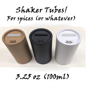 Puede incluir: Tres tubos cilíndricos para especias en color beige, negro y blanco. Cada tubo tiene una abertura ranurada y una serie de pequeños agujeros para dispensar. El texto dice "Shaker Tubes! For spices (or whatever)" y "3.25 oz (100ml)".