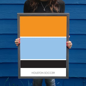 May include: Framed art print with a color block design in orange, white, light blue, black, and white. The words "HOUSTON SOCCER" are printed in white at the bottom of the print. The frame is dark gray.