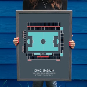 May include: A framed print of a soccer stadium, with a gray background and a teal field. The stadium is called "CPKC Stadium" and is located at 1460 E Front St Kansas City, Missouri. The coordinates are 39° 7' 50.88" N 94° 31' 29.856" W.
