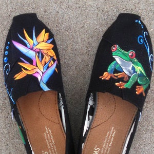 Puede incluir: Par de zapatos sin cordones TOMS negros, talla W6, con dise&ntilde;os pintados a mano. Un zapato presenta una colorida flor de ave del para&iacute;so, el otro una rana arb&oacute;rea verde. Ambos tienen plantillas marrones y el logo TOMS.