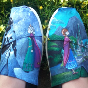 K&ouml;nnte beinhalten: Paar handbemalte Leinenschuhe mit Figuren aus dem Animationsfilm "Die Eisk&ouml;nigin". Ein Schuh zeigt eine Figur in einem gr&uuml;nen Kleid und lila Umhang vor blau-wei&szlig;em Hintergrund. Der andere Schuh zeigt eine Figur in einem blauen Kleid und lila Umhang.