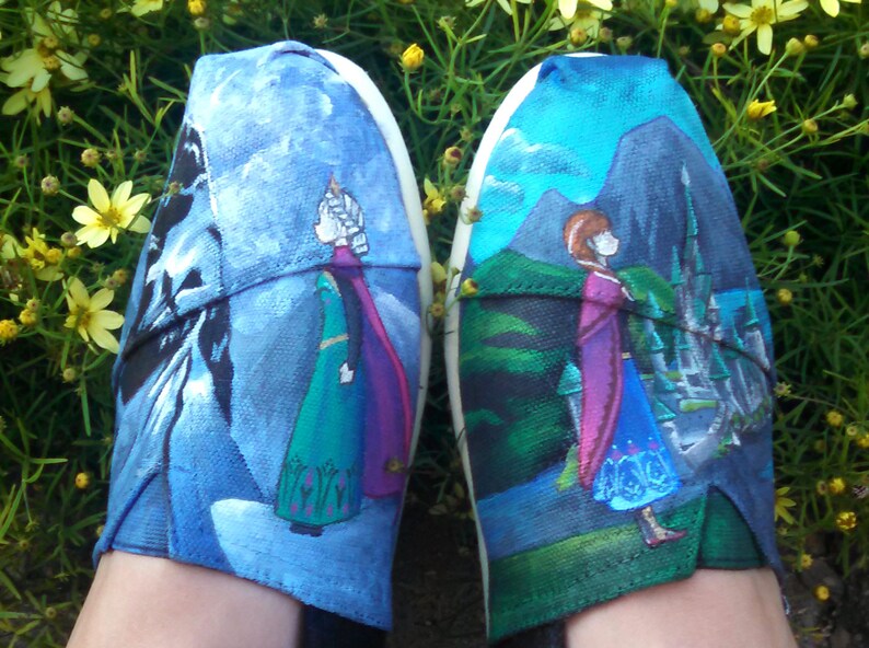 K&ouml;nnte beinhalten: Paar handbemalte Leinenschuhe mit Figuren aus dem Animationsfilm "Die Eisk&ouml;nigin". Ein Schuh zeigt eine Figur in einem gr&uuml;nen Kleid und lila Umhang vor blau-wei&szlig;em Hintergrund. Der andere Schuh zeigt eine Figur in einem blauen Kleid und lila Umhang.