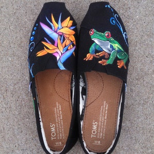 Puede incluir: Zapatos negros sin cordones con dise&ntilde;os pintados a mano. Uno presenta una colorida flor de ave del para&iacute;so, el otro una rana arb&oacute;rea verde con ojos rojos. Los zapatos son talla W6.