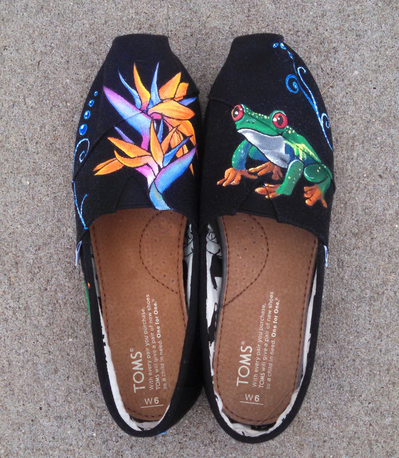 Puede incluir: Zapatos negros sin cordones con dise&ntilde;os pintados a mano. Uno presenta una colorida flor de ave del para&iacute;so, el otro una rana arb&oacute;rea verde con ojos rojos. Los zapatos son talla W6.