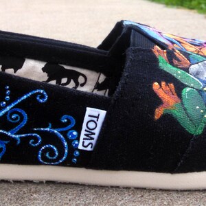 Puede incluir: Zapato negro sin cordones con suela blanca. El zapato presenta un dise&ntilde;o floral azul y una colorida ilustraci&oacute;n de una rana. El forro interior tiene un patr&oacute;n de silueta repetido. La marca "TOMS" es visible en el lateral.